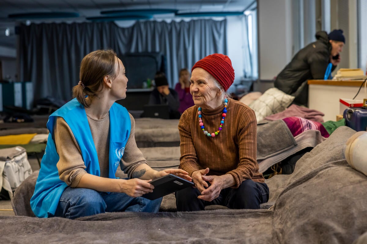 UNHCR staff member discussing with an older woman, both sitting on a bed.