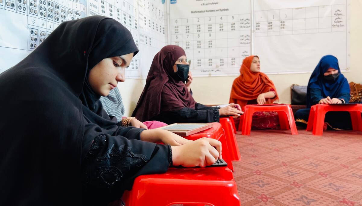 Helai Noori sits on the floor of a classroom with other women and reads Braille on a wooden tablet. Helai Noori sits on the floor of a classroom with other women and reads Braille on a wooden tablet.